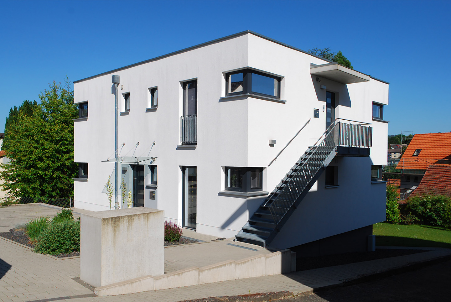 Realisiertes Gemeinschaftsprojekt der Architekturbüros stoffers architekten und Grohe und Budde Architekten, Inhaber Jochen Grohe, Diplom-Ingenieur, Architekt. Das Foto zeigt die Übereckansicht des Zweifamilienhauses mit strahlend weißer Putzfassade vor wolkenlosem, blauen Himmel. Das zweigeschossige Gebäude verfügt über zwei übereinanderliegende Wohnungen, die auf einem zurückspringenden, dunklen Betonsockel liegen. Über die Stahlaußentreppe gelangt man zur oberen Wohnung. Die klare Linienführung der Fenster und Außentüren mit anthrazitfarbenen Rahmen geben dem Gebäude ein modern klassisches Erscheinungsbild. Vor dem Haus sind links gepflasterte Stellplätze und ein grüner Vorgarten zu sehen. Über eine gepflasterte Rampe gelangt man in den Garten der Wohnanlage.