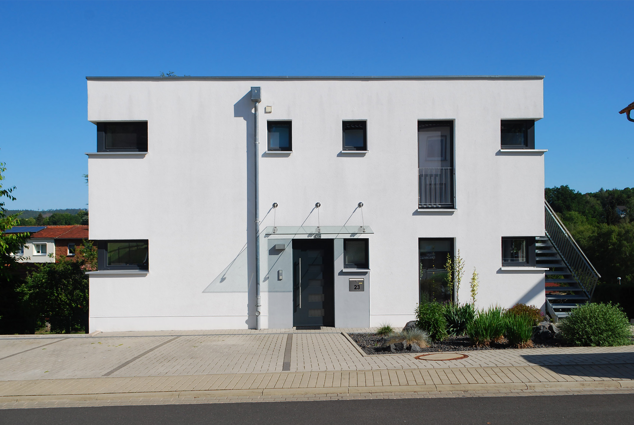 Realisiertes Gemeinschaftsprojekt der Architekturbüros stoffers architekten und Grohe und Budde Architekten, Inhaber Jochen Grohe, Diplom-Ingenieur, Architekt. Das Foto zeigt die Straßenansicht des Zweifamilienhauses mit strahlend weißer Putzfassade vor wolkenlosem, blauen Himmel. Das zweigeschossige Gebäude verfügt über zwei übereinanderliegende Wohnungen. Der Eingang der Erdgeschosswohnung befindet sich in zentraler Lage und besitzt ein abgehängtes Ganzglasvordach. Die Wohnung im Obergeschoss wird über eine Stahlaußentreppe auf der rechten Seite erschlossen. Die klare Linienführung der Fenster und Außentüren mit anthrazitfarbenen Rahmen geben dem Gebäude ein modern klassisches Erscheinungsbild. Vor dem Gebäude links befinden sich die gepflasterten Stellplätze. Ein gepflegter Vorgarten mit ausgewählter Bepflanzung rundet das harmonische Gesamtbild des Wohngebäudes ab.